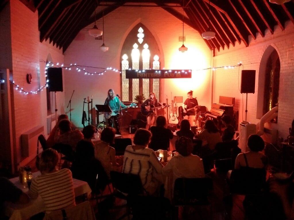 A group of people are gathered in a room lit with multi-coloured lights for a concert. The windows behind are arched glass and suggest this room was once a chapel.
