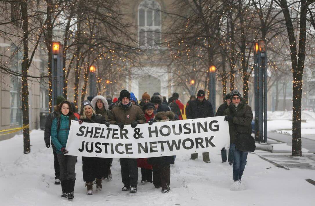 A group of people march through the snow with a banner which reads "Shelter and Housing Justice Network"
