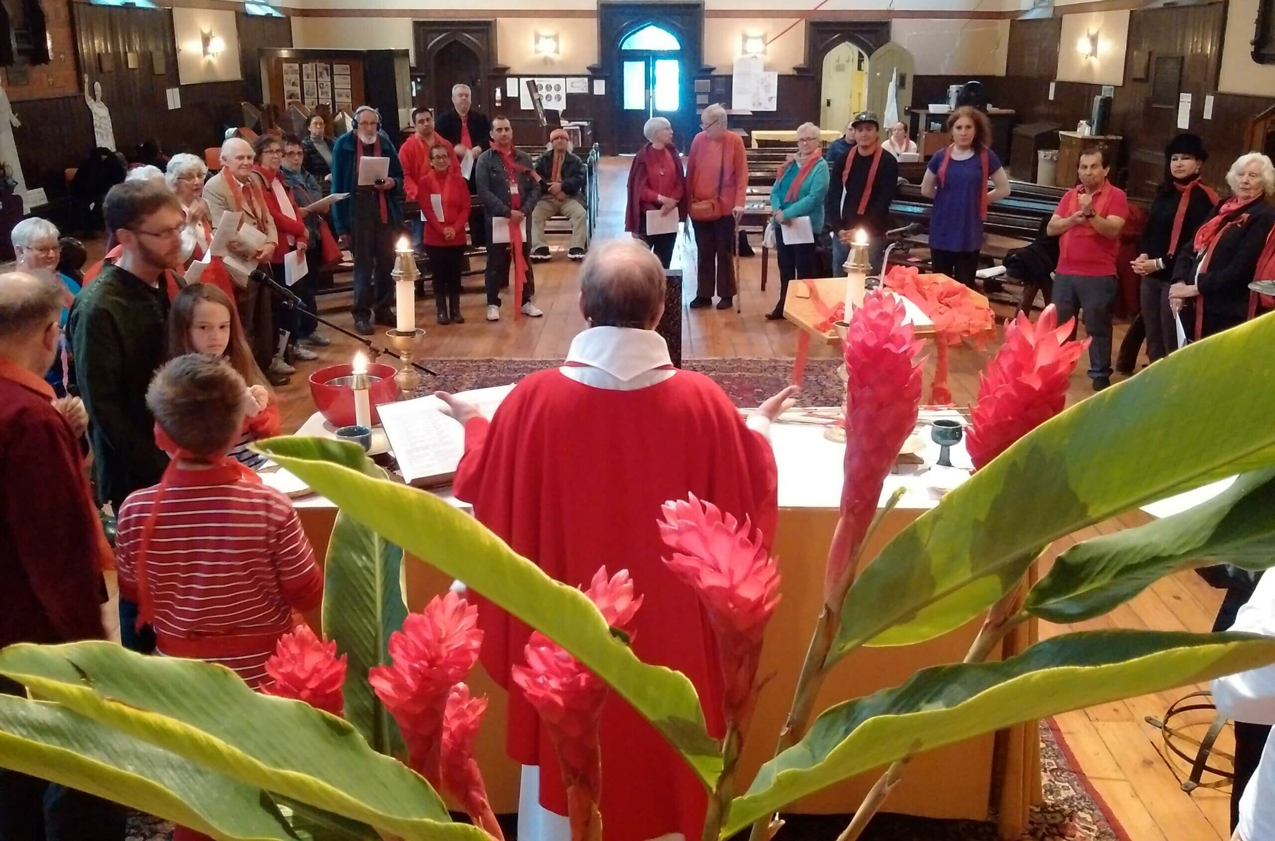 A man in a red cope has his back to the camera. He faces a congregation of people gathered to celebrate Pentecost. The image is taken through a bouquet of red flowers.